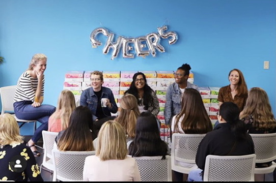Katy Simmons on a Girls Who Code panel, microphone in hand, facing a room of seated students with their hands raised