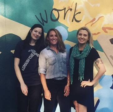 Three colleagues standing shoulder to shoulder in front of a large painted wall mural with the word work across it