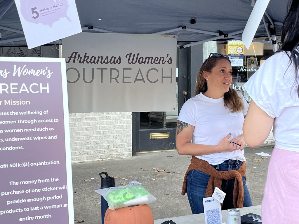 Katy Simmons leaning across an Arkansas Women's Outreach information table, mid conversation with a visitor picking up a flyer