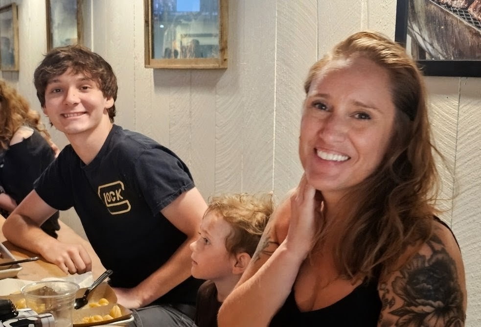 Katy Simmons seated at a restaurant table with her two sons, all leaning in for the photo